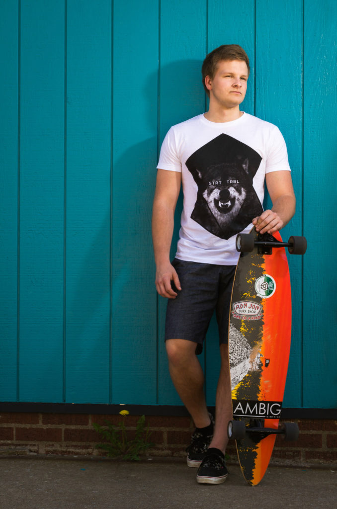 hipster guy leaning against blue wall holding skateboard