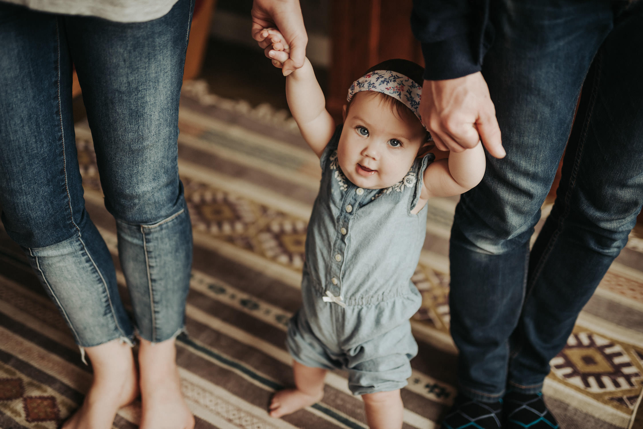 standing baby holding hands of parents while smiling at camera