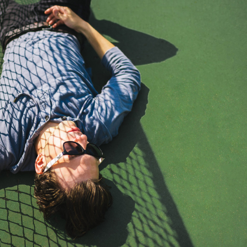 male model laying on green tennis court with shadows covering him