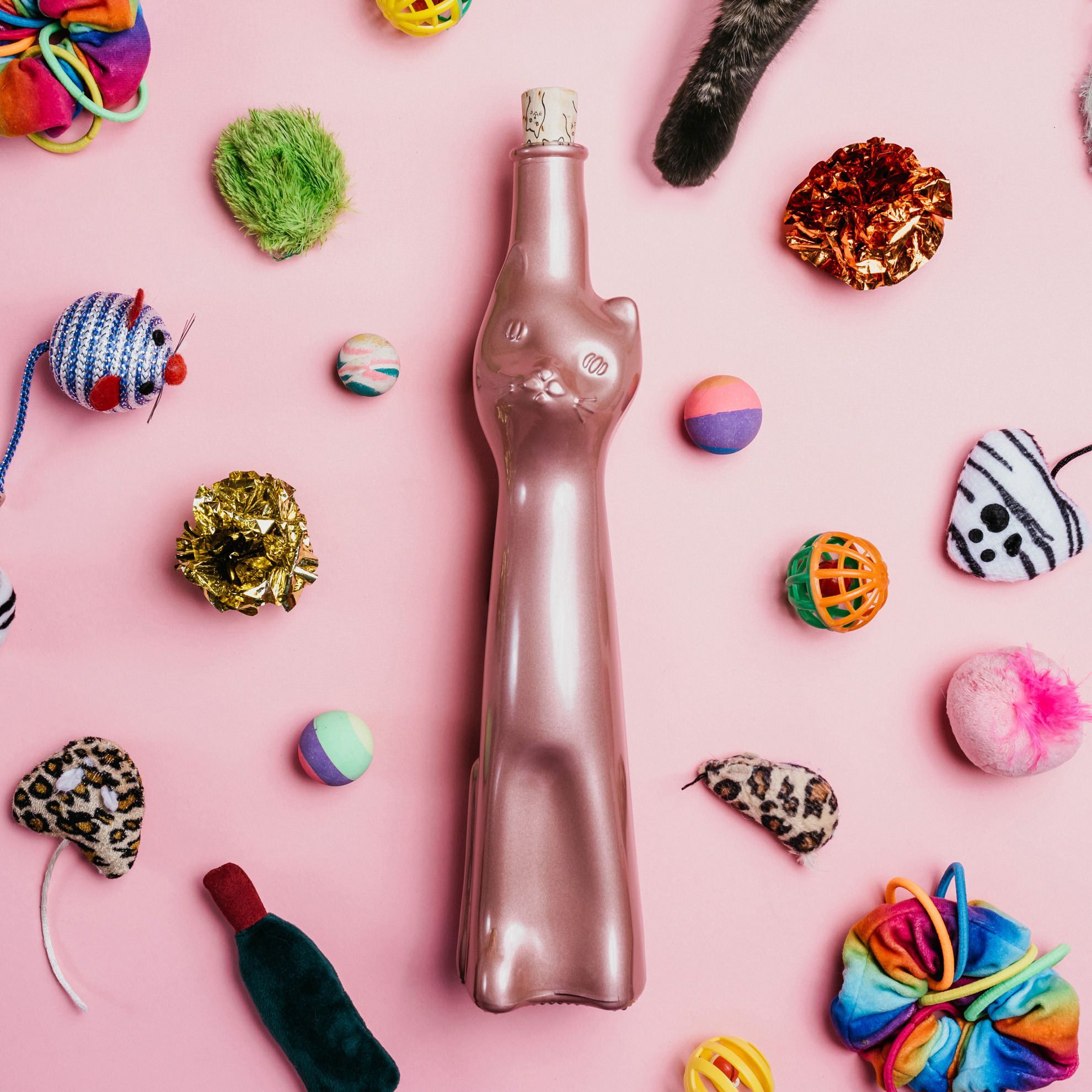 wine bottle in shape of cat laying on pink background surrounded by cat toys with a cat paw reaching towards it