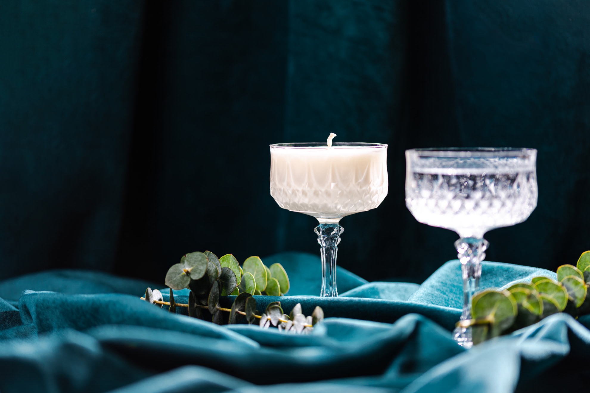 two vintage glasses with a cocktail in one glass and a candle in the other on a dark green velvet background